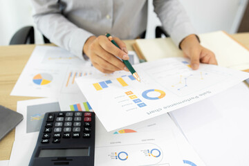 A woman is working on a desk in front of her with a pile of documents, a laptop, a calculator, a mobile phone and a notebook. This image shows her working on a project or analyzing data.
