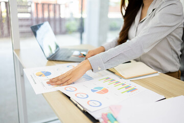 A woman is working on a desk in front of her with a pile of documents, a laptop, a calculator, a mobile phone and a notebook. This image shows her working on a project or analyzing data.
