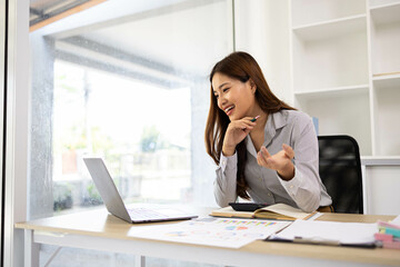 A woman is working on a desk in front of her with a pile of documents, a laptop, a calculator, a mobile phone and a notebook. This image shows her working on a project or analyzing data.
