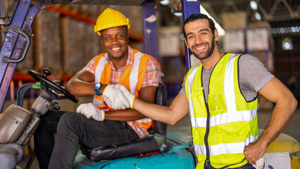 Fototapeta premium Two young male engineers in vest and hardhat shaking hands as a sign of peace gesture and agreement of deal and friendship in warehouse. african american worker sitting in forklift.
