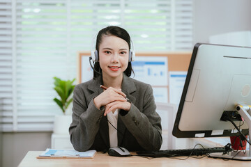 Smiling Asian female call center agent wearing a headset, speaking with a client at her office...