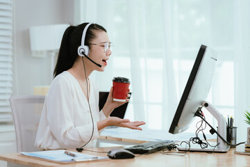Smiling Asian female call center agent wearing a headset, speaking with a client at her office...