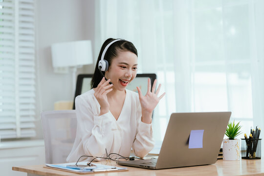 Smiling Asian female call center agent wearing a headset, speaking with a client at her office desk, providing professional customer support.