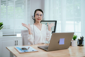 Smiling Asian female call center agent wearing a headset, speaking with a client at her office desk, providing professional customer support.