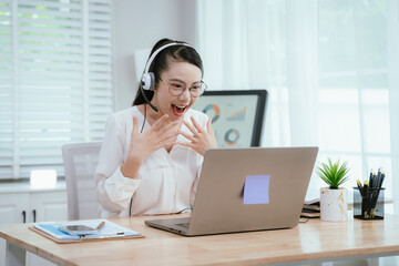 Smiling Asian female call center agent wearing a headset, speaking with a client at her office...