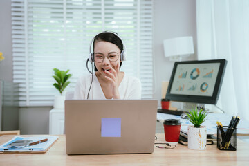 Smiling Asian female call center agent wearing a headset, speaking with a client at her office...