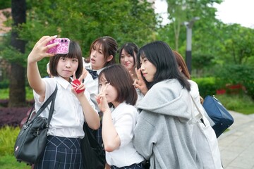 In early June in Pudong, Shanghai, seven Japanese high school girls in summer uniforms are taking a group selfie with a smartphone, surrounded by a lush green background near their school entrance.
