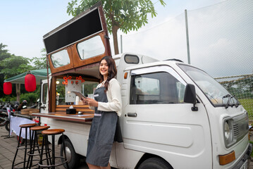 Indonesian southeast chinese asian female barista standing while holding a tablet in front of her coffee shop. A small business of a coffee shop or cafe on a truck