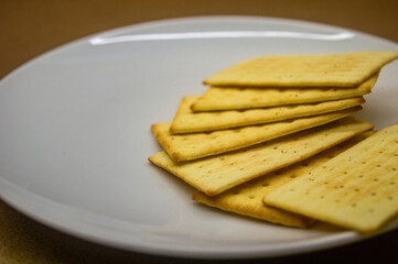 The stack of golden, rectangular crackers on a white plate, with a soft focus background. Perfect for food presentations, snack ideas, or culinary blogs