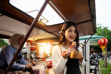 Indonesian southeast asian Chinese woman eating her food in front of a food truck with a Chinese muslim female waiter in a hijab inside it. A small business of a food truck