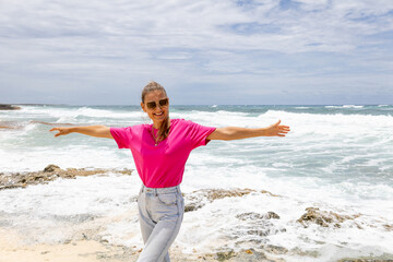 Cheerful woman in a bright pink t-shirt and light blue jeans enjoying the breeze on the rocky beach...