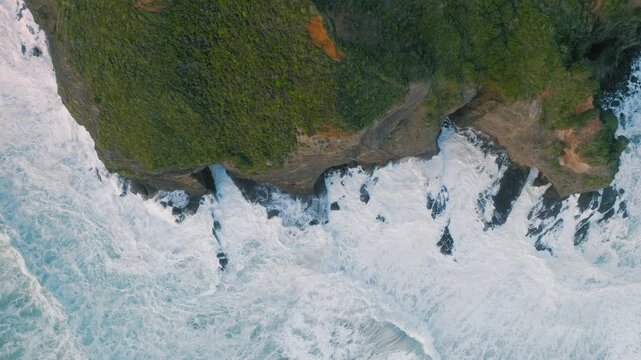 Aerial view of a waterfall cascading from a lush, green cliff into the turbulent ocean. The powerful waves crash against the rocky shore in New Zealand. Bethells Beach, NZ