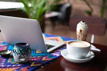 Photography of a work scene with a laptop, coffee, and ice cream dessert placed on a table with a colorful handmade tablecloth.