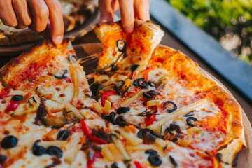 Men's hands tear off a slice of pizza with vegetable toppings, olives and stretching hot stretchy cheese. Process of eating pizza close-up. Delicious Italian pizza for friends and delivery