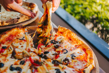 Men's hands tear off a slice of pizza with vegetable toppings, olives and stretching hot stretchy cheese. Process of eating pizza close-up. Delicious Italian pizza for friends and delivery