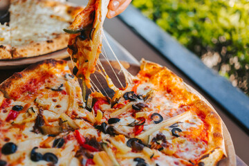 Men's hands tear off a slice of pizza with vegetable toppings, olives and stretching hot stretchy cheese. Process of eating pizza close-up. Delicious Italian pizza for friends and delivery