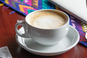 Photograph of a large coffee cup placed on a wooden table with a colorful textile tablecloth next to a computer