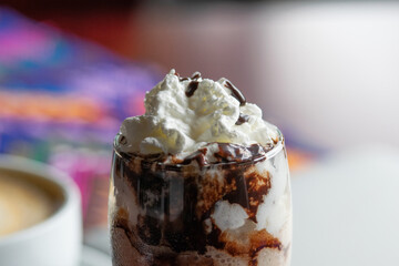 Photograph of a smoothie cocktail with chocolate ice cream in a glass with fresh whipped cream and a cup of coffee in the background