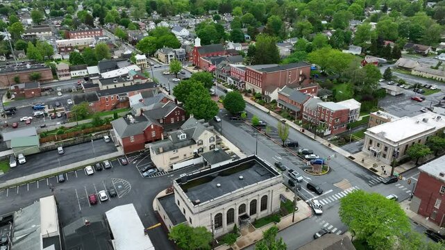 Establishing drone shot of american city with historic Buildings and flag of USA. Green trees during cloudy spring day. Descend aerial wide shot. Parking cars in center of city. Manheim,PA, USA.