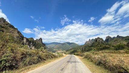Image depicts a scenic rural landscape featuring a paved road that stretches into the distance, flanked by lush, green vegetation on both sides