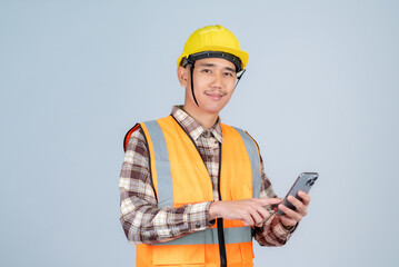 Smiling young Asian engineer or worker wearing yellow hard hat and reflective vest using smartphone working on grey background.