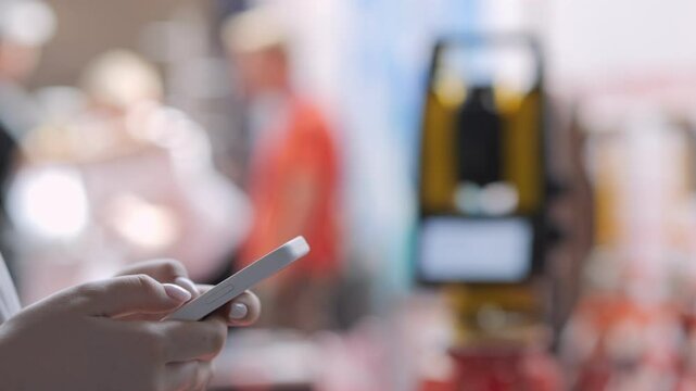 Person is holding a cell phone in a busy area. The background is blurry, and there are several people in the scene. Scene is busy and bustling, with people going about their daily activities