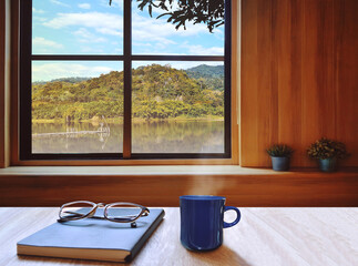 On the brown table, there are notebooks, books, diaries, blue coffee mugs and glasses. Outside the window, the atmosphere of nature is mountains, forests, trees, sky and clouds in Thailand. With copy 
