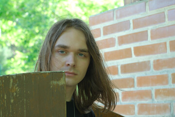 Young man with long hair poses near wooden structure in lush greenery