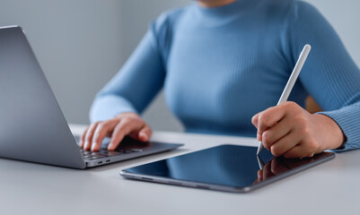 Close-up of businesswoman sitting at desk using stylus pen and digital tablet, working on laptop...