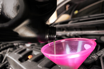 Engine oil or motor oil, is being poured from a dark bottle into a pink funnel in a car engine bay.