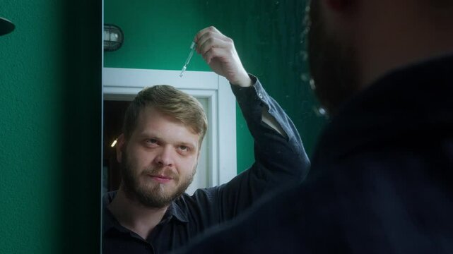 Young man looking in the mirror while using a dropper to apply hair serum to his scalp in a home bathroom setting.
