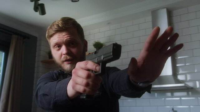 Serious man pointing a handgun with an extended arm in a modern kitchen, possibly acting in a dramatic or law enforcement role.
