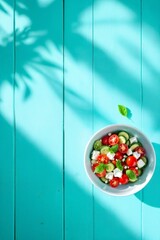 Fresh Greek salad in a white bowl placed on a bright turquoise wooden surface with leaf shadows. Perfect for healthy eating, summer recipes, or Mediterranean lifestyle content.