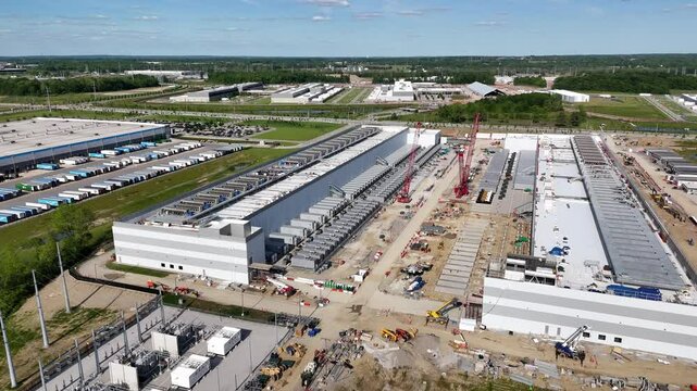 Drone footage of a massive industrial complex under construction with two large cranes operating between symmetrical rows of buildings. Dirt roads, materials, and active progress are clearly seen.