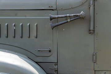 side of a military truck, showing a mounted dual trumpet horn. The olive green body features visible hinges and rivets, typical of vintage military vehicles