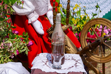 glass bottle with homemade alcohol placed on a doily, set against the background of a woman in traditional folk costume and a wooden spinning wheel. A scene from a fair or folk event, decorated with f