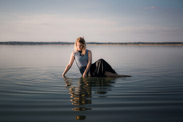 Fototapeta premium Young woman is sitting on a the beach in water and sunbathing. Rest on the sea. Girl portrait. Calmness. Happy face expression. Relaxed state. Dawn Meditation. Morning silence. Copy space. White skin.