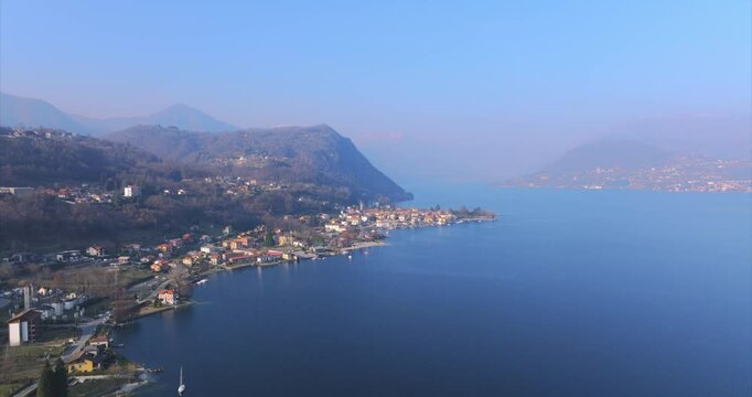 Aerial drone panoramic view of Pella village on picturesque shores of Lake Orta, Italy, hazy sunlight, mountains backdrop. Copy space