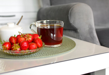 dojrzale truskawki i filiżanka herbaty na stoliku kawowym obok wygodnego fotela, zdrowy deser, relaks, ripe strawberries  and a cup of tea on a table next to a comfortable armchair, healthy desser
