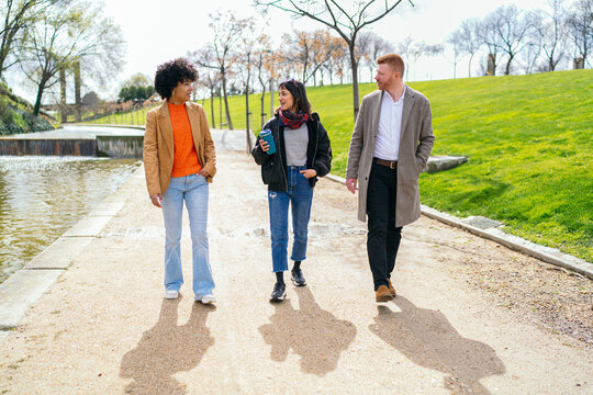 Three young professionals enjoying a conversation while walking in a city park next to a small pond, discussing business and enjoying the fresh air