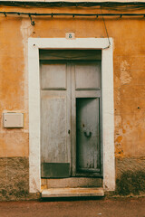 Weathered wooden door set in a textured orange wall. The door has a faded blue-gray hue and is framed by a white border. Above, a small plaque displays the number "8".