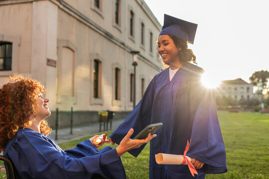 Two young women in graduation attire celebrating on a university campus, one in a wheelchair holding a diploma.