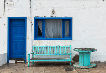 Weathered wooden bench with faded turquoise paint sits on a paved patio. Adjacent is a vivid blue door set in a white wall. A matching blue-framed window with net curtains is above the bench.