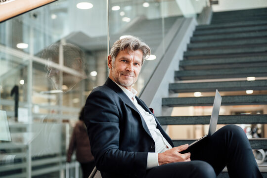 Confident businessman sitting on stairs with laptop in modern office
