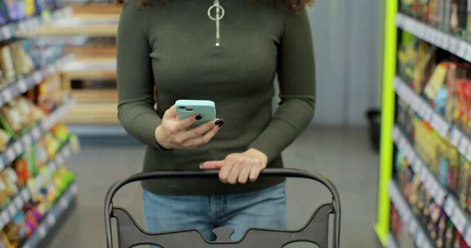 Close up of unrecognizable woman using smartphone while carrying shopping cart walking through aisles in hypermarket doing daily food shopping.