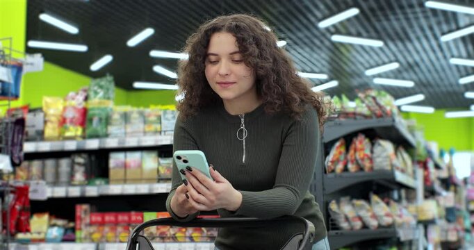 Beautiful woman use smartphone in supermarket. Woman buyer using mobile phone application standing with shopping trolley in supermarket.