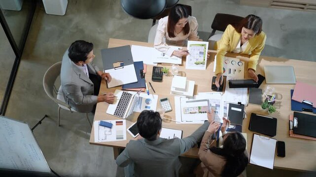 Productive corporate team discusses strategies and reviews printed charts around table during daytime meeting in home-based office setting.