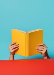 Hands holding a bright yellow book against a blue and red background in a creative indoor setting