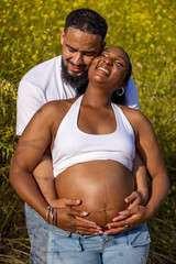 A loving couple enjoys summer outdoors, celebrating their pregnancy. The husband hugs his wifes baby bump, showing their deep connection and love, surrounded by vibrant flowers and greenery
