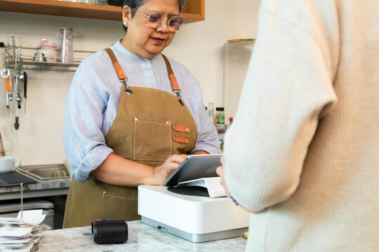 Senior entrepreneur cafe owner barista processes credit card payment on terminal, Asian female in apron smiling toward client across tiled counter, secure cashless sales inside modern coffee shop
